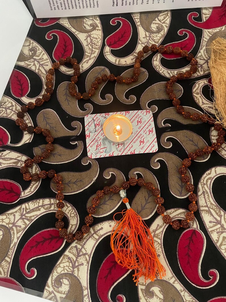 May include: A brown beaded mala necklace with an orange tassel, set on a patterned surface with red and brown paisley designs. A lit candle and a small card are also visible.