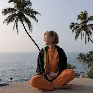 May include: A person in orange pants and a long-sleeved orange shirt sits cross-legged on a concrete surface, looking to the side. They wear a black shawl with green trim and a beaded necklace. Palm trees and the ocean are in the background.