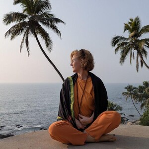 May include: A person in orange trousers and a long-sleeved orange top sits cross-legged on a concrete surface, looking to the side. They wear a black shawl with green trim and a beaded necklace. Palm trees and the ocean are in the background.