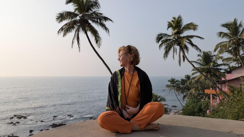 May include: A person in orange trousers and a long-sleeved orange top sits cross-legged on a concrete surface, looking to the side. They wear a black shawl with green trim and a beaded necklace. Palm trees and the ocean are in the background.