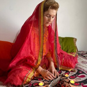 May include: A person wearing a red head covering and orange clothing, seated on a patterned surface. Lit candles, a metal feather, and prayer beads are arranged in front of them. The scene evokes a sense of meditation or ritual.