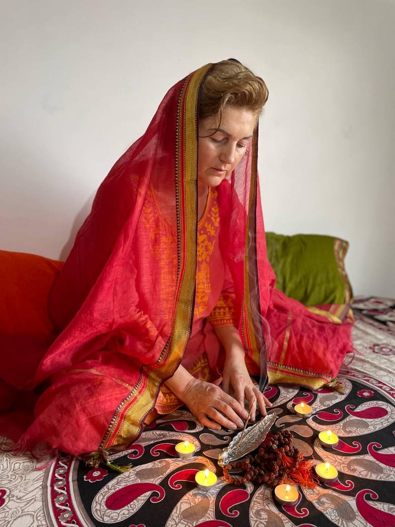 May include: A person wearing a red head covering and orange clothing, seated on a patterned surface. Lit candles, a metal feather, and prayer beads are arranged in front of them. The scene evokes a sense of meditation or ritual.