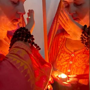 May include: An image of a person wearing a red and orange patterned garment, holding a lit candle. The person is wearing a beaded bracelet. The scene is illuminated with warm, reddish light.