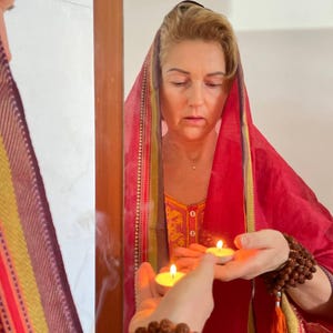 May include: A woman in a red headscarf and dress, holding two lit candles. The woman is reflected in a mirror. A strand of brown beads is wrapped around her wrist. The scene suggests a spiritual or ritualistic practice.