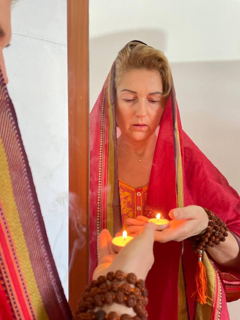 May include: A woman in a red headscarf and dress, holding two lit candles. The woman is reflected in a mirror. A strand of brown beads is wrapped around her wrist. The scene suggests a spiritual or ritualistic practice.