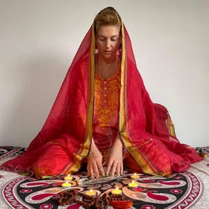 May include: A person in a red and gold embroidered outfit and head covering, seated on a patterned blanket. Lit candles and incense are arranged in front, suggesting a spiritual or meditative practice.