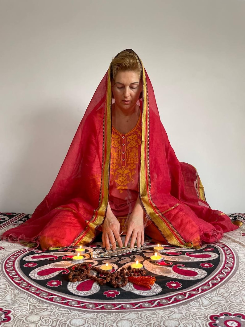 May include: A person in a red and gold embroidered outfit and head covering, seated on a patterned blanket. Lit candles and incense are arranged in front, suggesting a spiritual or meditative practice.
