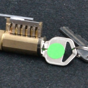 May include: A brass key cylinder with a silver key and a green tag attached. The key cylinder has a series of pins visible.