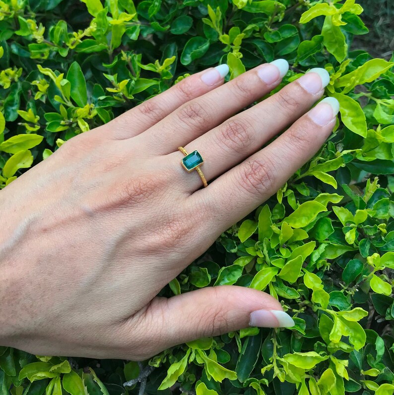 以下が含まれることがあります： 長方形のエメラルドカットが施された緑色の宝石をあしらったゴールドの指輪。シンプルな金属製のバンドは、表面にざらざらとした質感の加工が施されており、宝石は4本の爪で固定されています。
