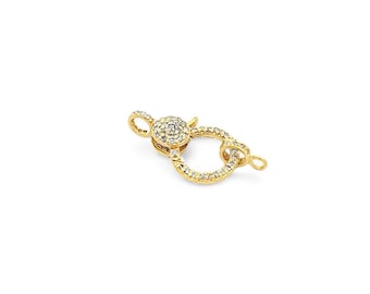 May include: A gold-toned circular jewelry clasp featuring a spring-loaded mechanism and a pavé-set surface adorned with numerous small, clear, sparkling stones. The clasp includes two small loops on either side for attachment to jewelry chains.