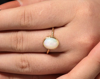 Può includere: Un anello in oro con una pietra preziosa di opale bianco di forma ovale al centro, caratterizzata da riflessi iridescenti. L'incastonatura presenta una finitura testurizzata dall'aspetto organico e il sottile gambo è anch'esso in oro testurizzato.