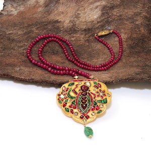May include: A gold pendant necklace with a detailed design featuring a ruby-encrusted insect, surrounded by green and red gemstones. The necklace is strung with red ruby beads.