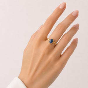 以下が含まれることがあります： ベゼルセッティングにセットされた滑らかな楕円形の青い宝石が特徴のゴールドリング。リングのバンド部分はテクスチャーのあるわずかにハンマーで叩いたような仕上げになっており、中央の青い石の磨かれた表面と繊細なコントラストを生み出しています。