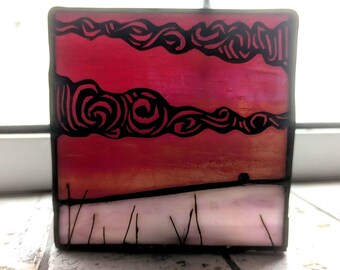 May include: A square stained glass panel showing a sunset. The sky is a gradient of red and orange, with black swirling cloud-like shapes. The bottom features a white and pink landscape with black vertical lines.