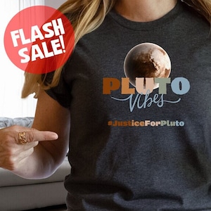 Könnte beinhalten: Ein dunkelgraues T-Shirt mit einer Grafik von Pluto und dem Text "Pluto Vibes" und "#JusticeForPluto". Die Grafik ist in Braun-, Orange-, Blau- und Weißtönen gehalten. Das T-Shirt wird von einer Person mit langen blonden Haaren getragen.