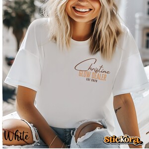 May include: White t-shirt with brown text that reads "Christine Glow Dealer Est 2023".