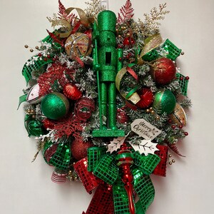 May include: A green glitter Christmas wreath with a green glitter nutcracker, red and green ornaments, gold ribbon, and a sign that says "Merry & Bright Christmas".