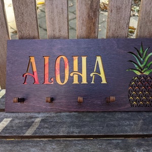 May include: Wooden key holder with the word "ALOHA" in a rainbow gradient and a pineapple design. The key holder has four hooks.