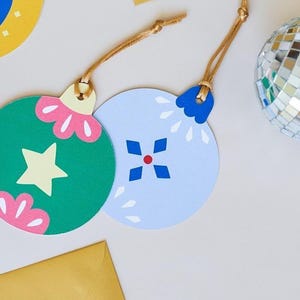 Op de afbeelding: Een verzameling kleurrijke, ronde kerstversieringen met verschillende ontwerpen, waaronder een boom, een ster en een maan. De ornamenten zijn blauw, groen en geel, met gouden touwhangers. Twee discoballen en gele enveloppen zijn ook aanwezig.