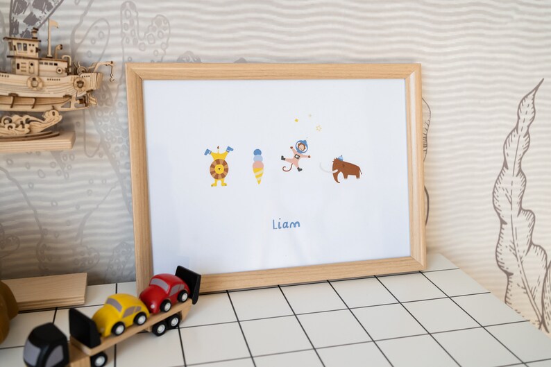 May include: A framed print with a white background featuring five cartoon characters: a monster, an ice cream cone, an astronaut, a mammoth, and a star. The name "Liam" is printed below the characters.