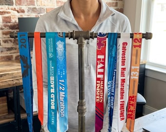 Puede incluir: Un soporte de exhibición de medallas de metal con varias medallas de finalista de maratón y cintas coloridas. Las cintas tienen texto como "1/2 Marathon", "HALF MARATHON FINISHER" y "13.1". El soporte está sobre una superficie de madera.