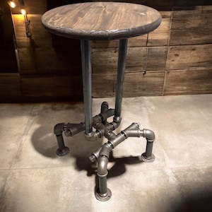 May include: A rustic, industrial-style side table. It features a round, stained wooden top supported by two vertical metal pipes. The base is constructed from interconnected metal pipes and fittings, forming a unique, three-legged design. The table is suitable for indoor use.