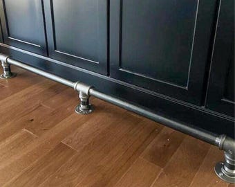 Puede incluir: Un reposapiés metálico, construido con tuberías y accesorios industriales, está montado en un armario oscuro. El riel está soportado por bridas circulares y se coloca sobre un suelo de madera. El diseño tiene una estética rústica e industrial.