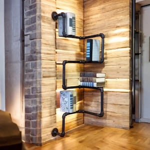 May include: A black pipe shelving unit mounted on a wooden wall, holding several books. The industrial-style shelves are arranged in a zig-zag pattern. The wall is made of light-colored wooden planks, and the floor is a light brown wood.