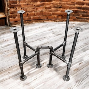 May include: Industrial-style table legs made of dark gray metal pipes and fittings. The legs have a sturdy design with circular mounting plates at the top and bottom, connected by a network of pipes. The legs are designed to be attached to a tabletop.