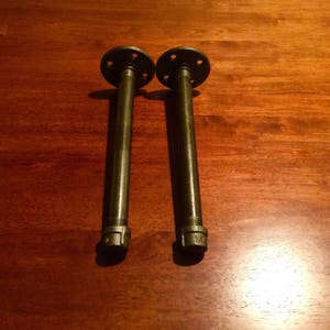 May include: Two black metal pipe shelf brackets with flanges. The brackets are about 10 inches long.