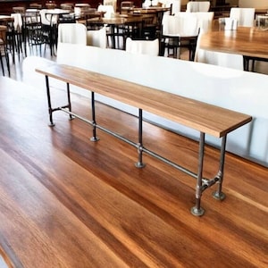 May include: A long wooden bench with a light brown finish. The bench is supported by black metal legs and a horizontal pipe. The bench is in a restaurant setting with other tables and chairs visible in the background.
