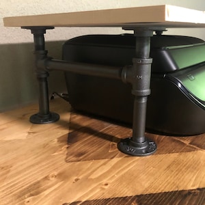 May include: A rustic printer stand made from industrial pipes and a light-colored wooden shelf. The stand features black metal pipes and fittings, with a rectangular wooden top. The stand is designed to elevate a printer.