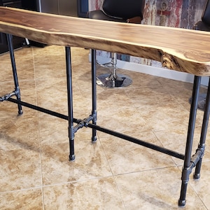 May include: A long, rectangular wooden table with a live edge. The tabletop is a rich brown color with a lighter edge. The table is supported by black metal pipe legs and crossbars, creating an industrial aesthetic.