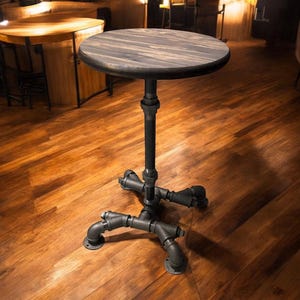 May include: A round, dark-stained wooden table with a black metal pipe base. The table is approximately 30 inches in diameter and stands on a four-legged base. The industrial design is suitable for a bar or cafe setting.
