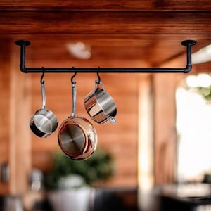 May include: A black metal pot rack suspended from a wooden ceiling. Three stainless steel pots and a copper-bottomed pan hang from the rack. The pots are different sizes and shapes, with shiny surfaces. The rack is a practical kitchen storage solution.