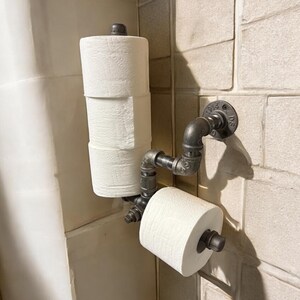 May include: Industrial-style toilet paper holder made from galvanized steel pipes. The holder is mounted on a tiled wall and holds two rolls of white toilet paper. The design features a unique, rustic aesthetic.