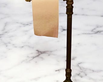 Puede incluir: Un portarrollos de papel higiénico de estilo industrial. Está hecho de tuberías y accesorios de metal oscuro, con una base redonda y un brazo horizontal para sostener el rollo. El rollo de papel es de color marrón claro.