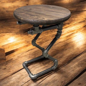 May include: A small, industrial-style side table with a round, dark-stained wooden top. The base is constructed from black metal pipes and fittings, creating a unique, geometric design. The table is approximately 18 inches tall.