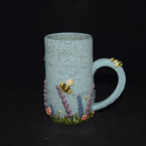 May include: A tall, light blue speckled ceramic mug featuring a sculpted design of green grass, purple and blue lavender sprigs, and small yellow and black bees. One bee is positioned on the handle, while others are on the mug body.