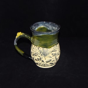 May include: A ceramic mug with a green and blue glazed exterior. The handle has a yellow floral accent. The lower portion of the mug features a black and white illustrated design with the words "Smoking Area".