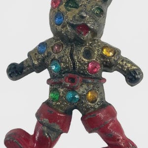 May include: A vintage cat-shaped brooch with a metallic finish, adorned with colorful rhinestones. The cat figure is wearing a red belt and boots. The brooch has a whimsical, antique aesthetic.