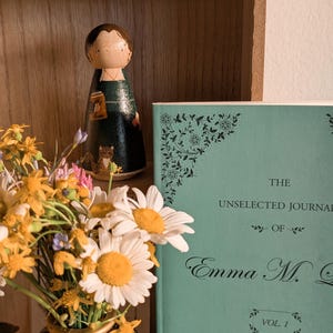May include: A wooden peg doll with a dark green dress and a book in hand stands on a wooden shelf. A book with a teal cover and the title "The Unselected Journal of Emma M. G." is next to the doll. A vase of daisies and wildflowers is in the foreground.