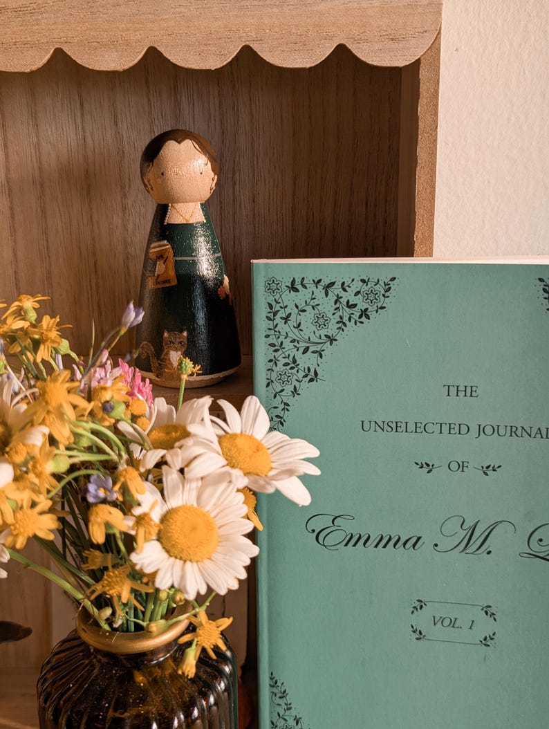 May include: A wooden peg doll with a dark green dress and a book in hand stands on a wooden shelf. A book with a teal cover and the title "The Unselected Journal of Emma M. G." is next to the doll. A vase of daisies and wildflowers is in the foreground.