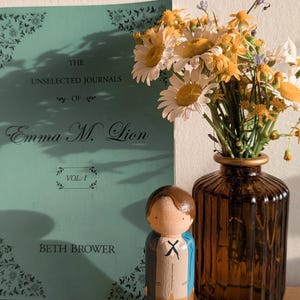 May include: A still life featuring a teal book titled "The Unselected Journals of Emma M. Lion" with a wooden peg doll and a vase of daisies. The doll is painted with a blue jacket and tan pants. The vase is brown.