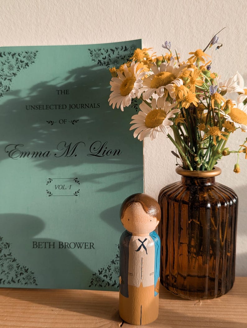 May include: A still life featuring a teal book titled "The Unselected Journals of Emma M. Lion" with a wooden peg doll and a vase of daisies. The doll is painted with a blue jacket and tan pants. The vase is brown.