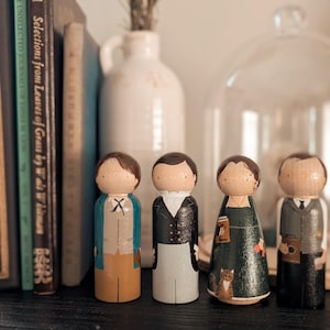 May include: Four wooden peg dolls, each with painted details depicting figures in various outfits. The dolls stand in front of a shelf with books and a glass cloche. The dolls have brown hair and are dressed in different styles.