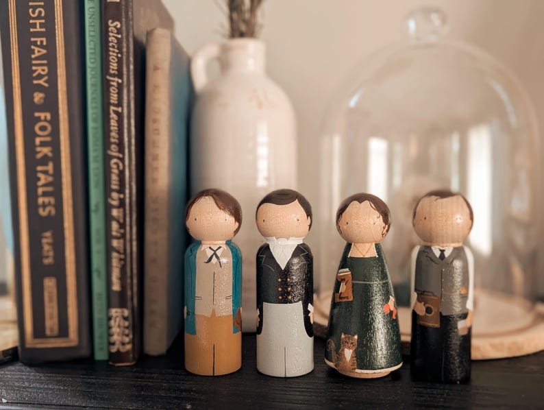 May include: Four wooden peg dolls, each with painted details depicting figures in various outfits. The dolls stand in front of a shelf with books and a glass cloche. The dolls have brown hair and are dressed in different styles.