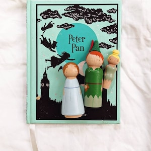 May include: A vintage Peter Pan book with a green cover and a black and white illustration of Peter Pan flying. Three wooden peg dolls are placed on top of the book. The dolls are dressed as Peter Pan, Wendy, and Tinkerbell.