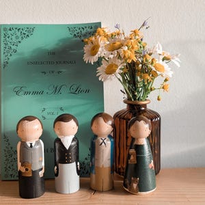 May include: Wooden peg dolls dressed in vintage-style clothing stand in front of a book and a vase of flowers. The book is titled "The Unselected Journals of Emma M. Lion." The dolls are painted with details.