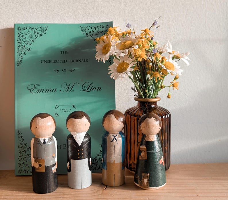 May include: Wooden peg dolls dressed in vintage-style clothing stand in front of a book and a vase of flowers. The book is titled "The Unselected Journals of Emma M. Lion." The dolls are painted with details.